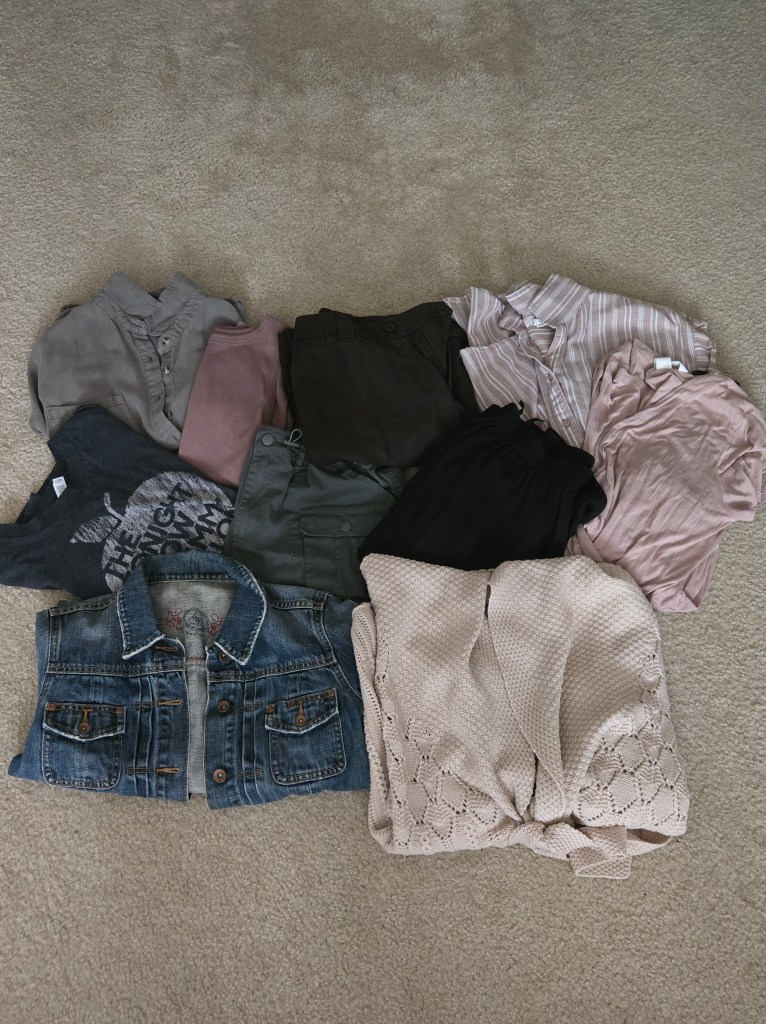 A collection of ten versatile clothing pieces arranged on a beige carpet, including shirts, trousers, a denim jacket, and a cardigan, showcasing a muted earth-tone palette.