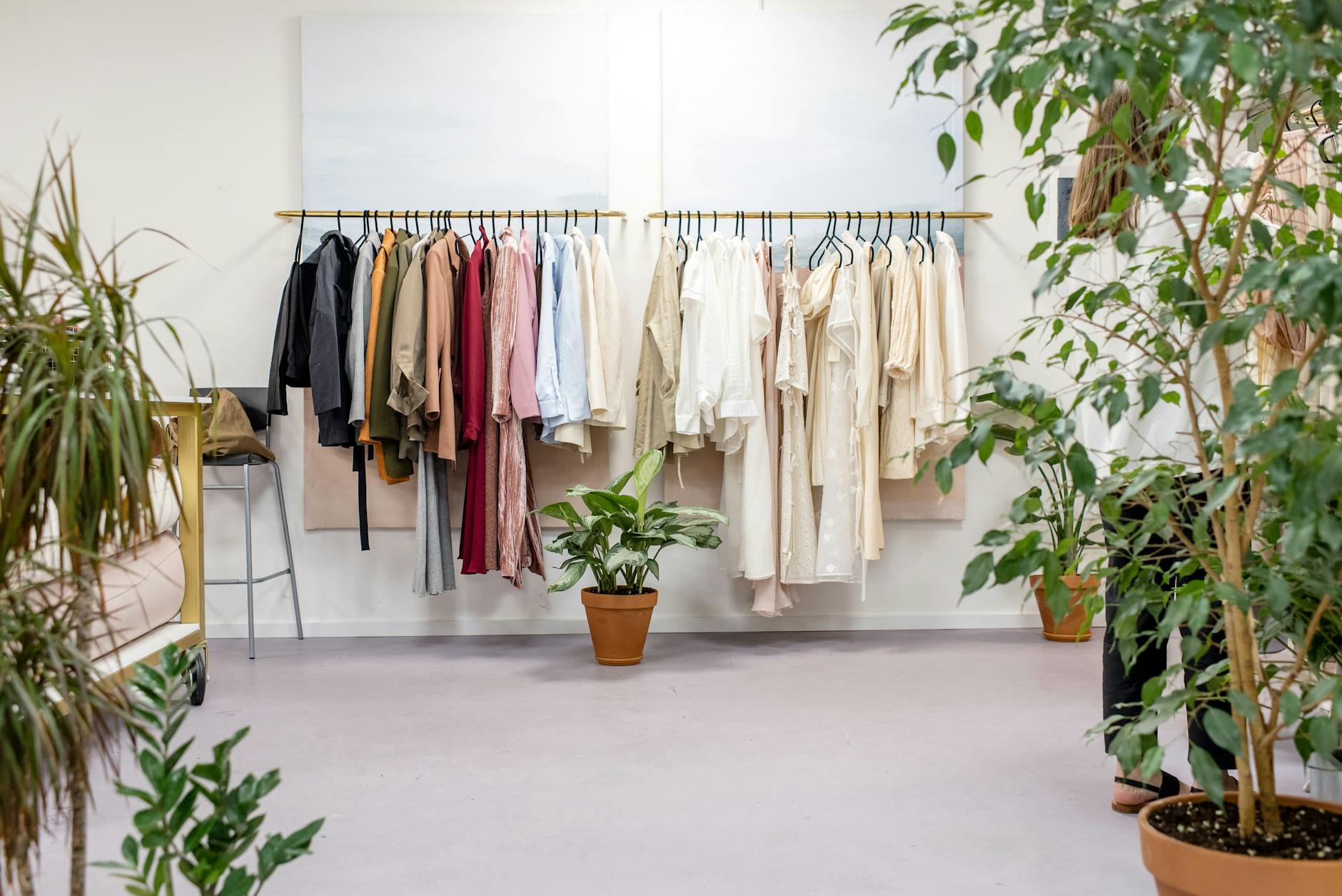 A well-organized clothing rack displaying a variety of tops in neutral and earth tones, surrounded by indoor plants in a bright, cozy room.